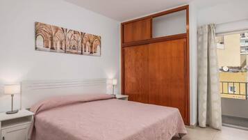 A bedroom with a bed, bedside tables, a wooden wardrobe, a painting on the wall, and a view through a window.