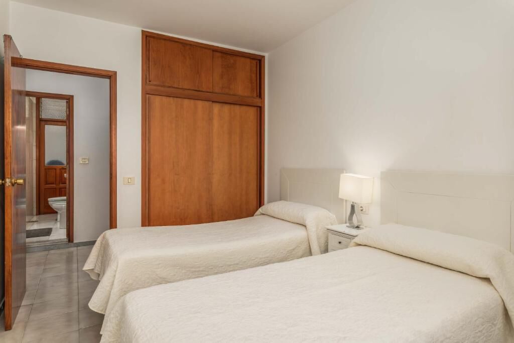 A hotel room with two single beds, a wooden wardrobe, a bedside table with a lamp, and a bathroom visible through an open door.