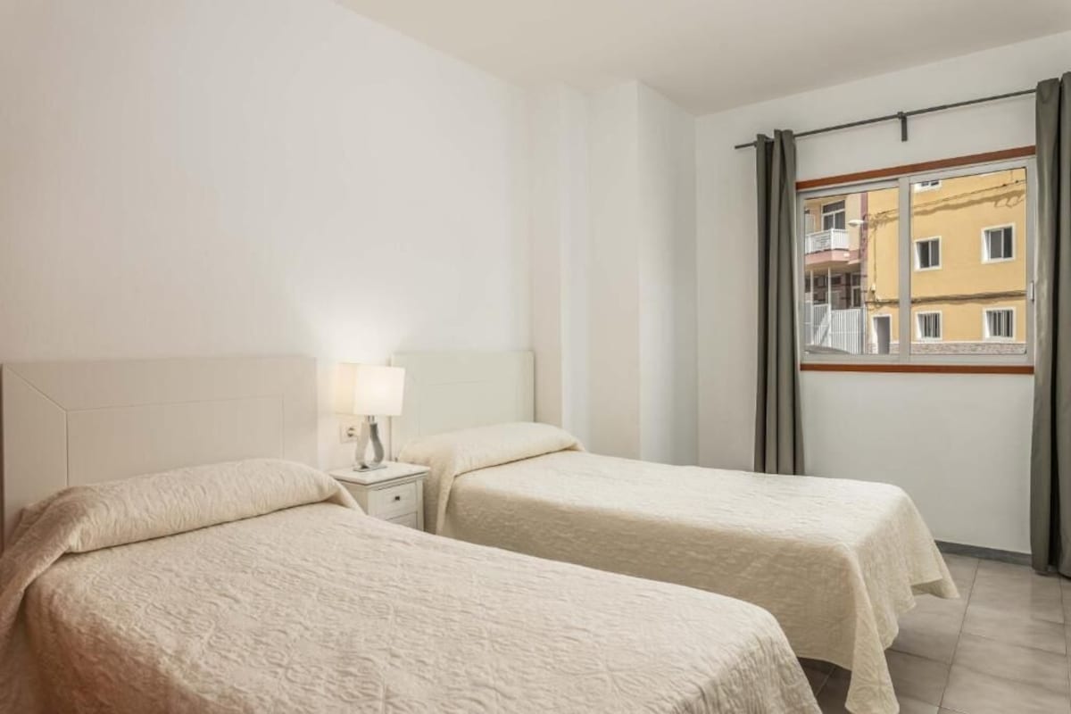 A hotel room with two beds, a nightstand with a lamp, and a window with a view of buildings.