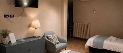 Habitación de hotel con televisión de pantalla plana, escritorio con lámpara, silla, cama con manta azul y radiador.
