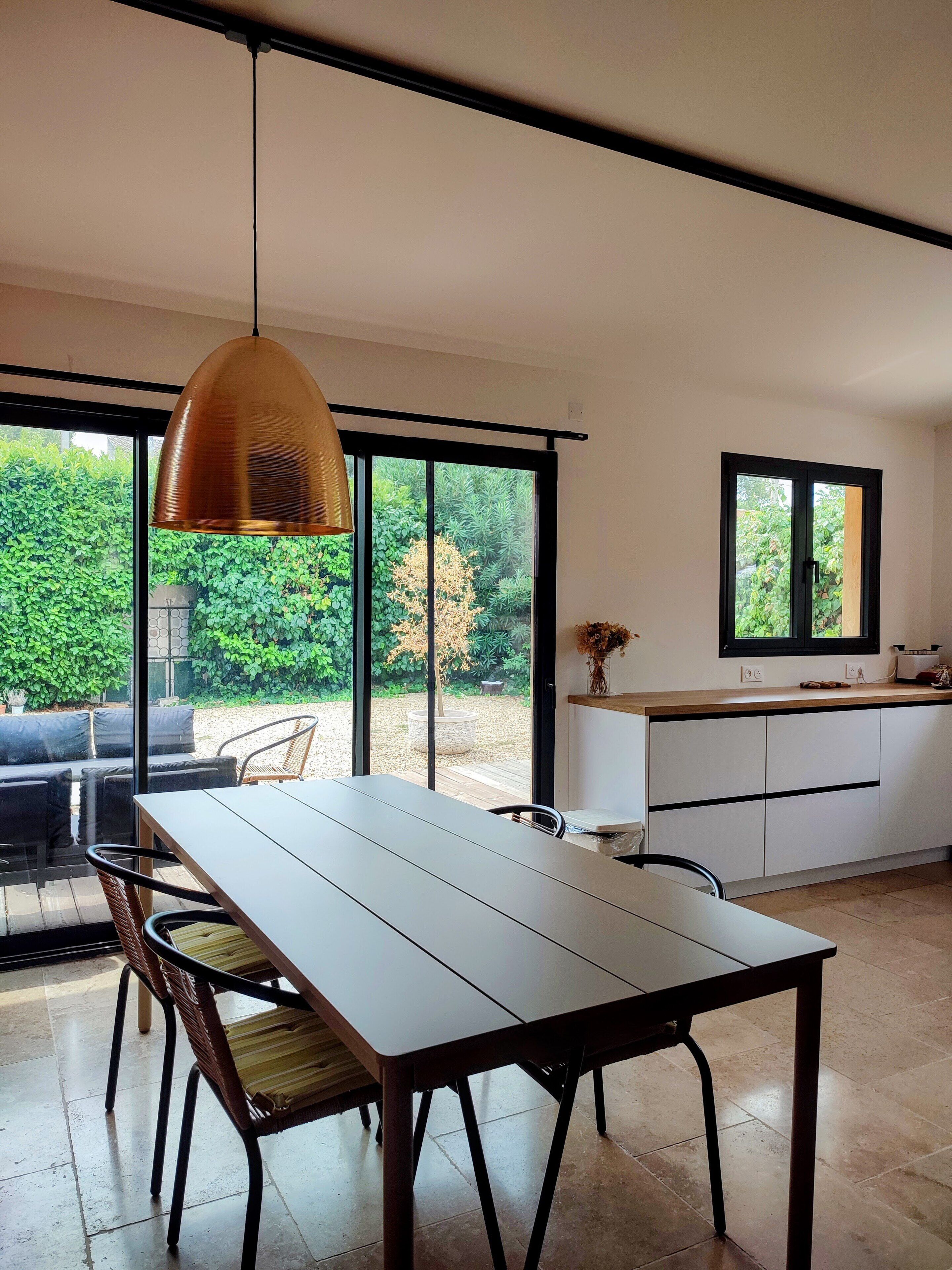 A modern kitchen with a dining area, large windows, and a view of greenery.