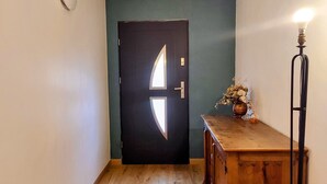 Un pasillo con piso de madera, una puerta oscura con una lámpara y una pequeña mesa con un jarrón de flores secas.