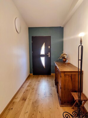 A hallway with a wooden floor, a dark door with a light fixture, and a small table with a vase of dried flowers. - A charming house in earthy tones with an intimate garden where you can relax (Avignon)