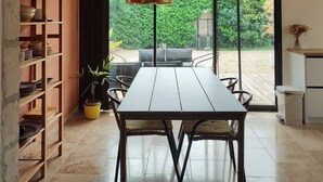 Un comedor moderno con una mesa y sillas, una lámpara colgante y vistas a un jardín a través de puertas de cristal grandes.