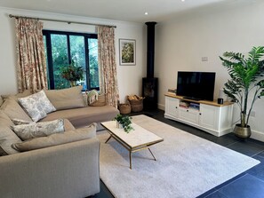 A living room with a sofa, a coffee table, a TV on a cabinet, a fireplace, and a window with curtains. - Beautiful Cotswold Cottage | Sleeps 6-8 | Near to Elkstone Studios (Winstone)