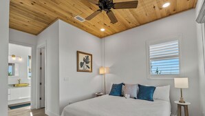 Ein Schlafzimmer mit einem Bett, einem Deckenventilator, einem Fenster mit Jalousien, einer Nachttischlampe und einem Wandbild.