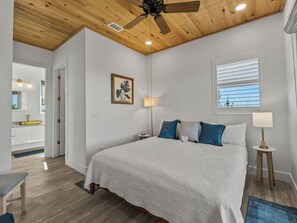 Ein Schlafzimmer mit einem Bett, einem Deckenventilator, einem Fenster mit Jalousien, einer Nachttischlampe und einem Wandbild.