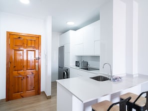 A modern kitchen with white cabinetry, a wooden door, stainless steel appliances, and a bar area with chairs. - Toboso 2J Torrecilla Beach by Casasol by Interhome (Nerja)