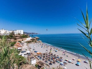 Une plage côtière avec un ciel dégagé, un parachute ascensionnel visible au loin, ainsi qu’une variété de parasols et de chaises longues.
