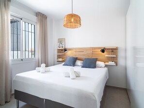 Quarto de hotel com cama, mesa de cabeceira, luminária, janela com cortinas e prateleira na parede.