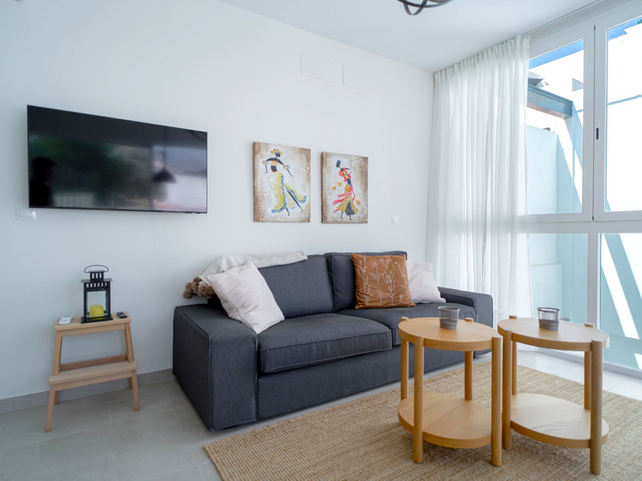 Un salon moderne avec un canapé gris, deux tables d’appoint, un téléviseur à écran plat et des œuvres d’art accrochées au mur.