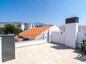 Um terraço no telhado com soalho em mosaico, uma mesa com cadeiras e vista para edifícios e montanhas ao fundo.