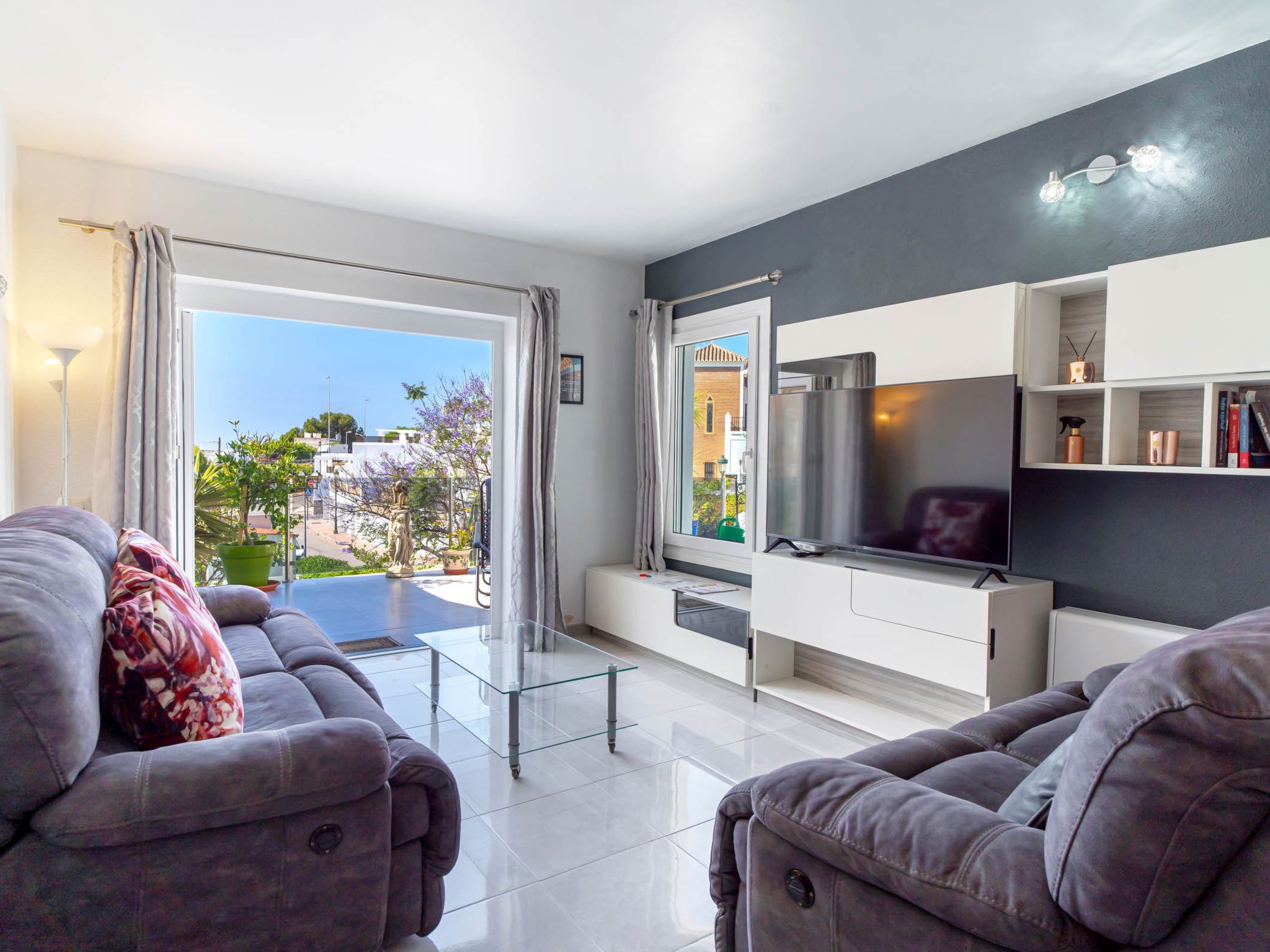 Ein modernes Wohnzimmer mit einer Sofagarnitur, einem Flachbildfernseher und einem Balkon mit Blick auf Gebäude und Grünflächen.