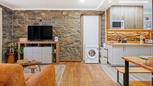 Ett kompakt bostadsutrymme med en stenmur, en platt-TV, en tvättmaskin och ett litet kök.