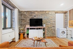 A living room with a stone wall, a flat-screen TV, a washing machine, and a small table with a book. - Stone House Rochinha by Homie (Funchal)