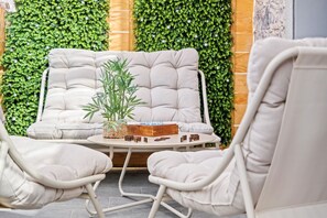 Eine Sitzecke mit gepolstertem Sofa und zwei Sesseln, einem kleinen Tisch mit Pflanze und Dominosteinen sowie einer grünen, blätterreichen Wand im Hintergrund.