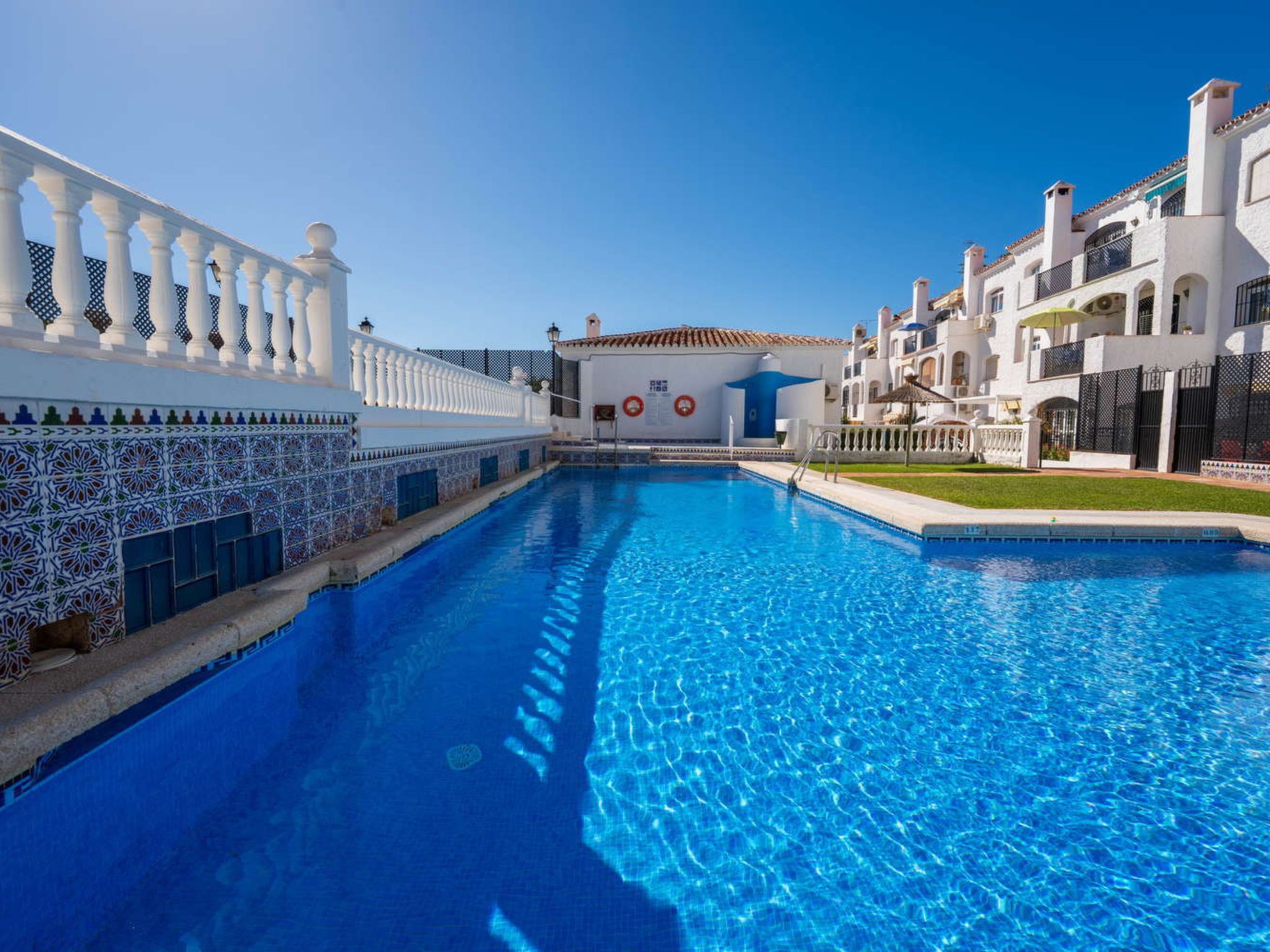 Una piscina con superficie de agua azul cristalina, rodeada de edificios blancos con balcones y un sendero con baldosas.