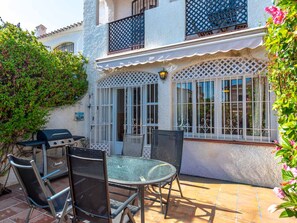 Eine Terrasse mit einem runden Glastisch, Metallstühlen, einem Grill und einem weißen Gebäude mit Gitterfenstern.