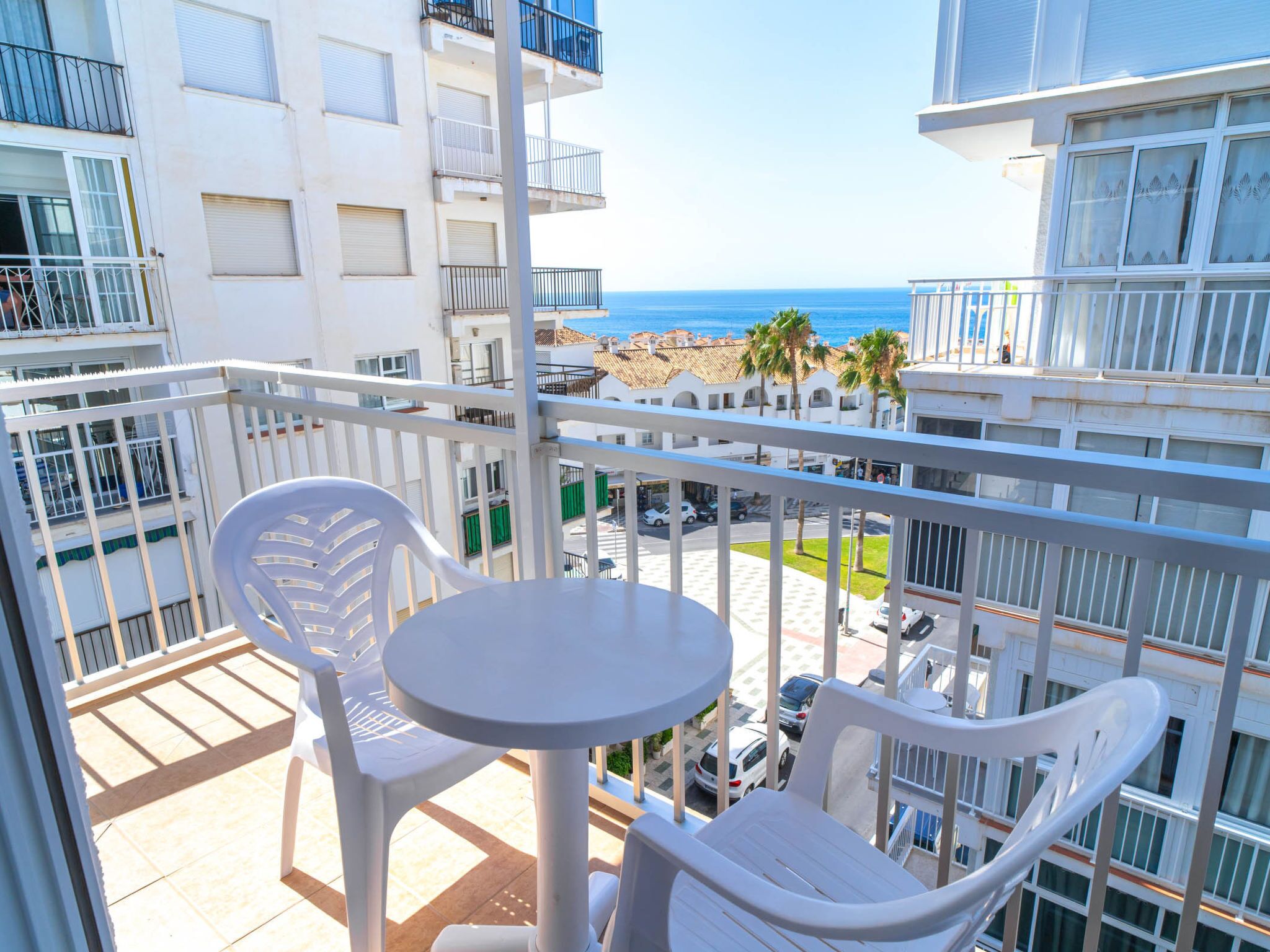 Ein Balkon mit weißen Plastikstühlen und einem kleinen Tisch, der auf eine Küstenlandschaft mit Gebäuden und Palmen blickt.