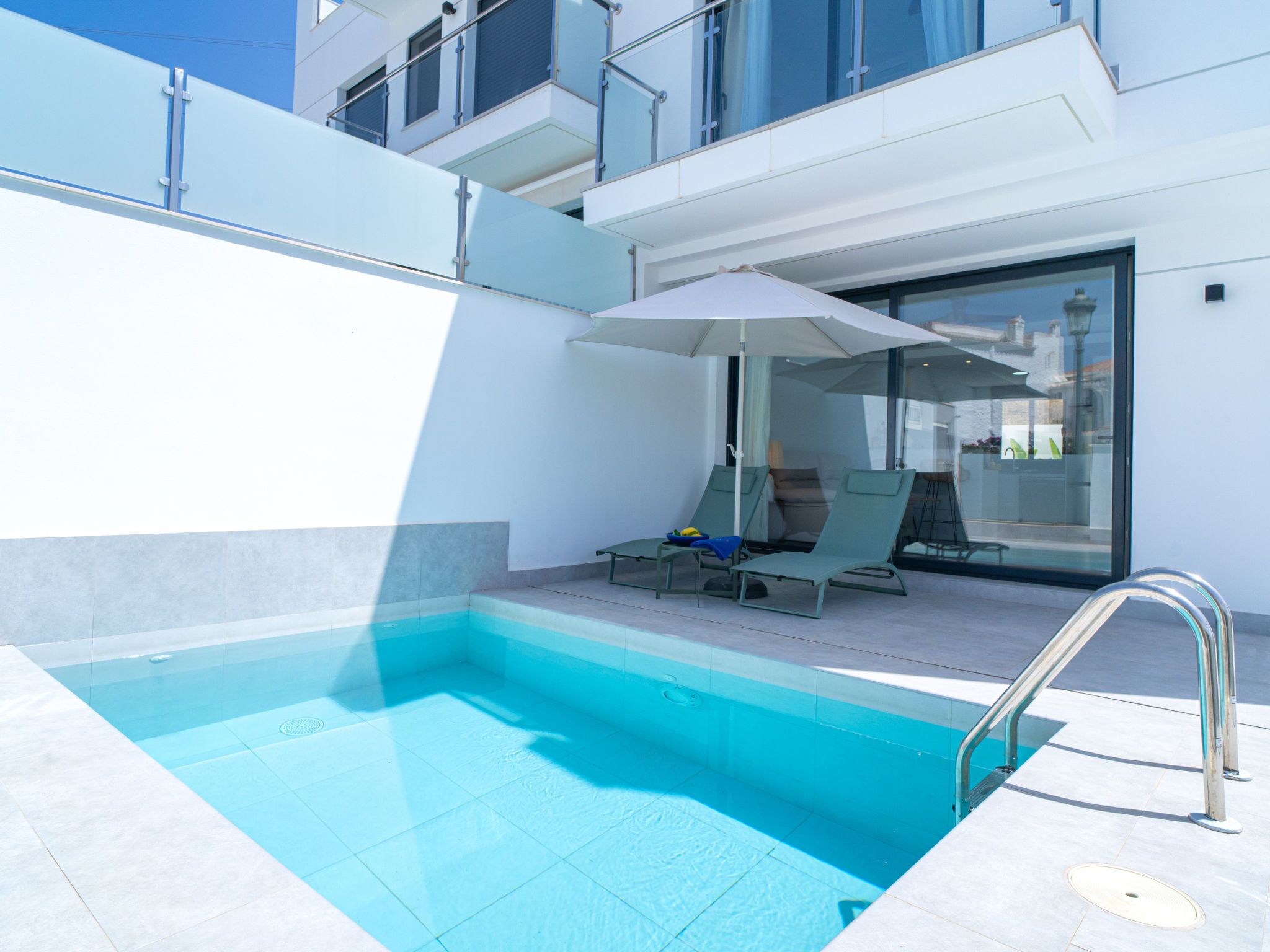 A modern outdoor pool area with a glass balustrade, two lounge chairs, and an umbrella.