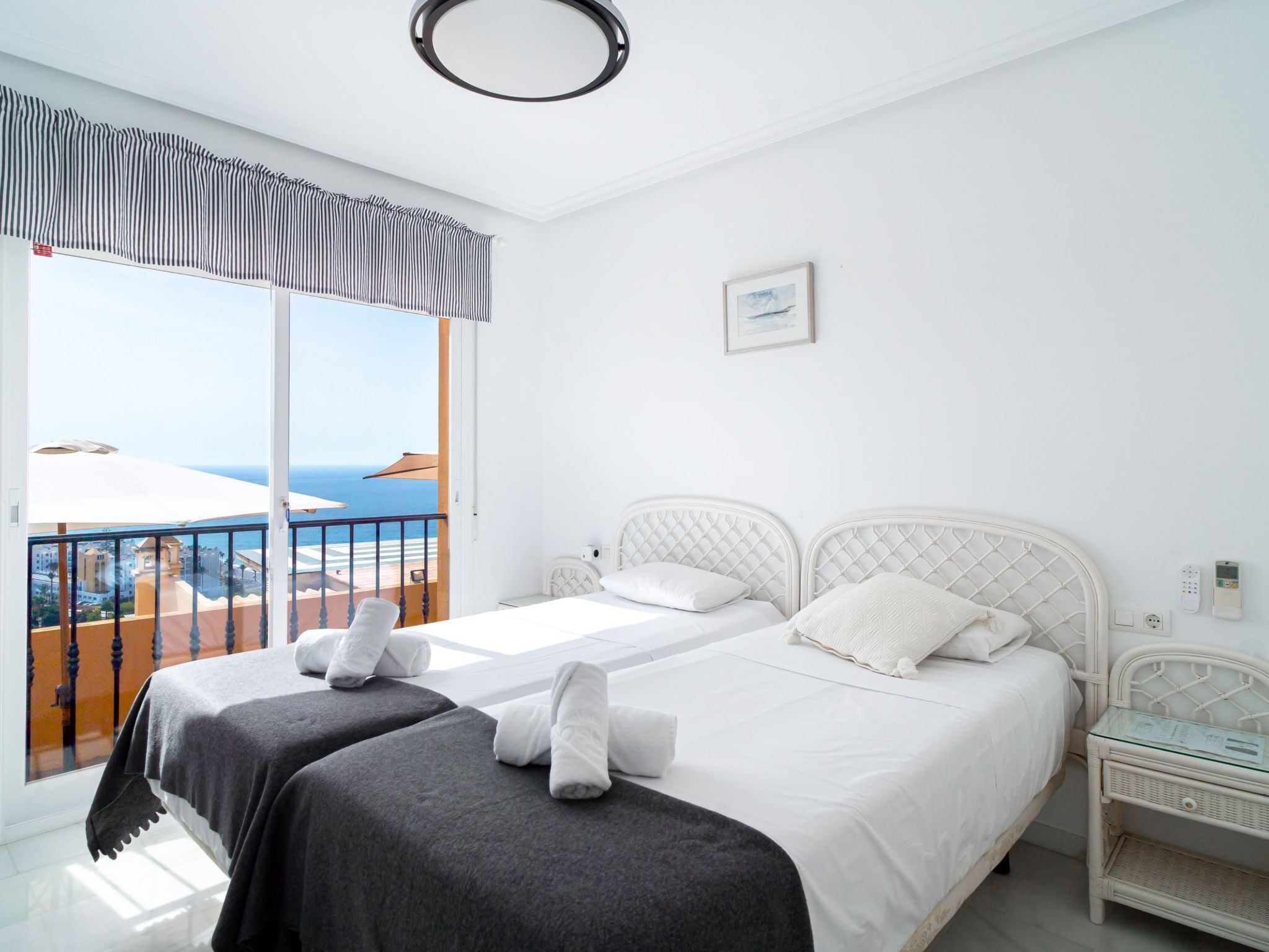 A hotel room with two beds, a balcony with a view of the sea, and a small bedside table with a lamp.