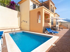 Una piscina amb aigua transparent de color blau, envoltada per un solàrium de rajoles, i un edifici amb un balcó i mobiliari exterior.