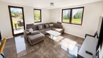 A modern living room with a sectional sofa, a coffee table, and large windows offering views of a green landscape.