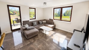A modern living room with a sectional sofa, a coffee table, and large windows offering views of a green landscape.