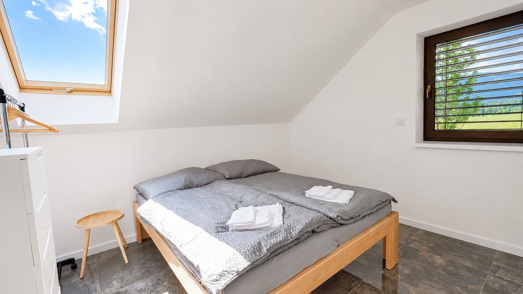 A bedroom with a bed, a small stool, a window with blinds, and a view of greenery outside.