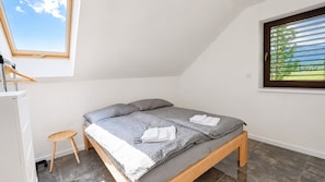 A bedroom with a bed, a small stool, a window with blinds, and a view of greenery outside.