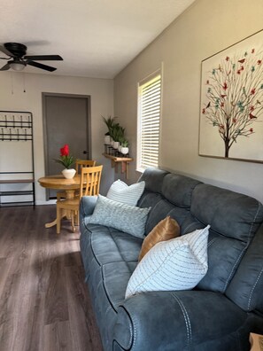 A living room with a dark grey sofa, wooden dining table, and chairs. There is a ceiling fan, a window with blinds, and a wall-mounted shelf with plants. - NeNe's House (Russellville)