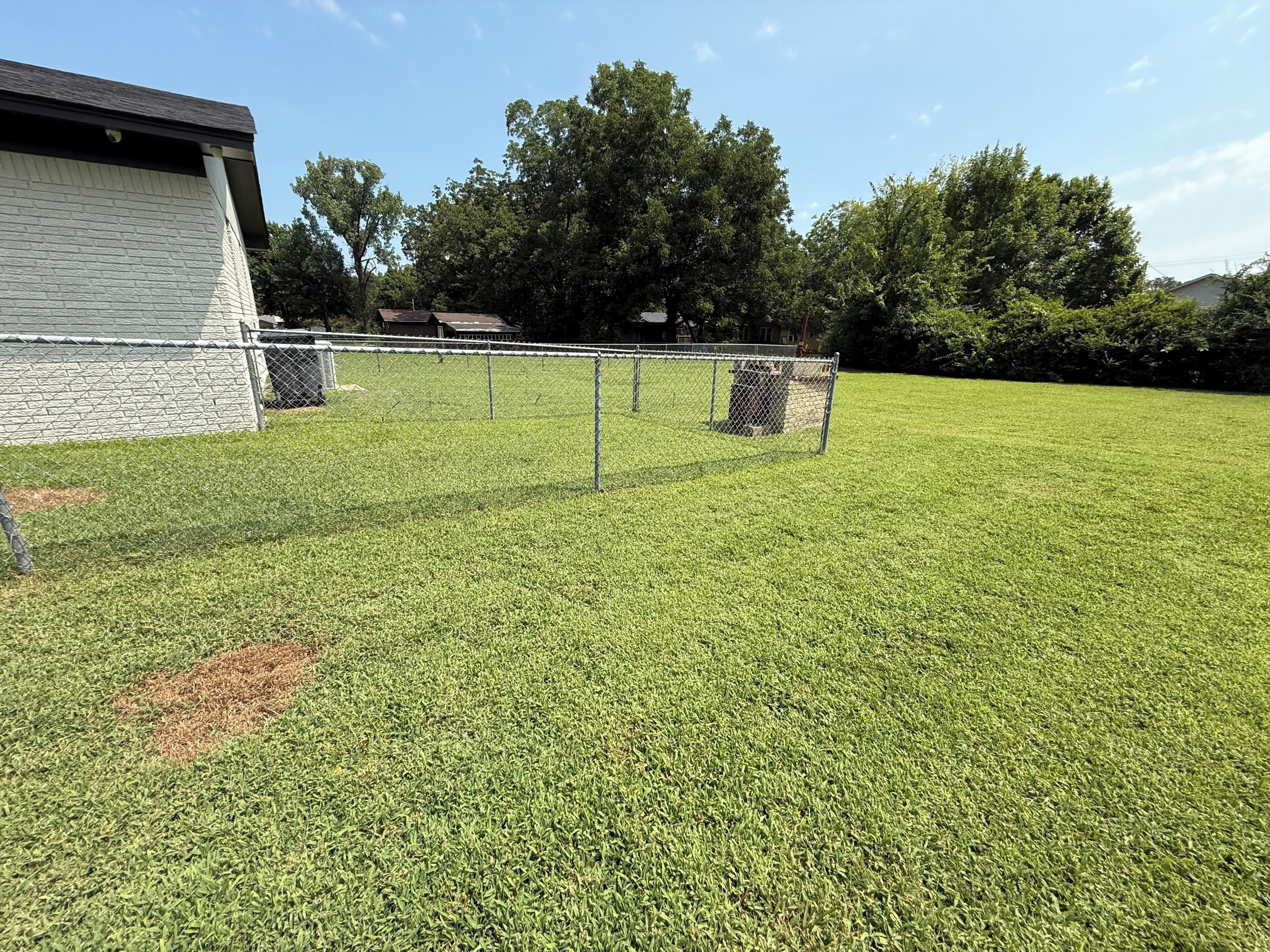A fenced yard with a grassy area, a building with a white exterior, and trees in the background.