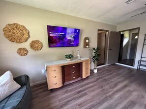 A living room with a flat-screen TV mounted on the wall, a wooden console table with drawers, a potted plant, and a decorative wall hanging. - NeNe's House (Russellville)