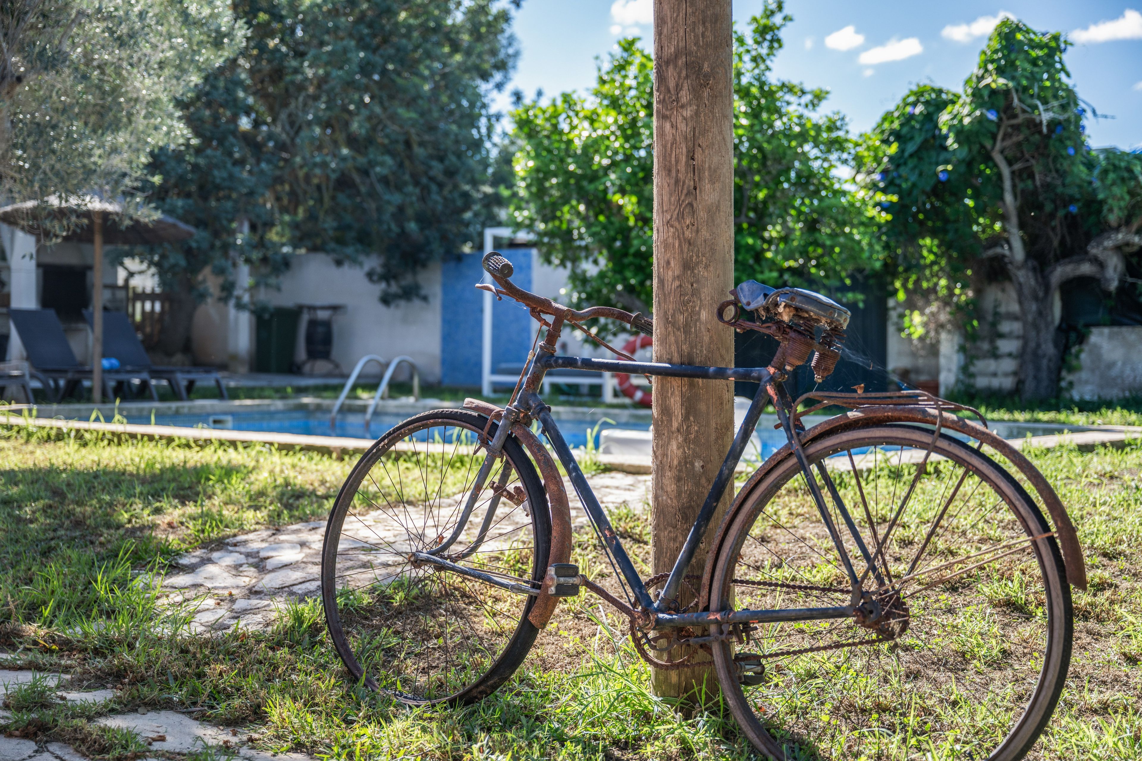 Una bicicleta estacionada junto a un poste de madera en un área con césped, con una piscina y sillas de descanso al fondo.