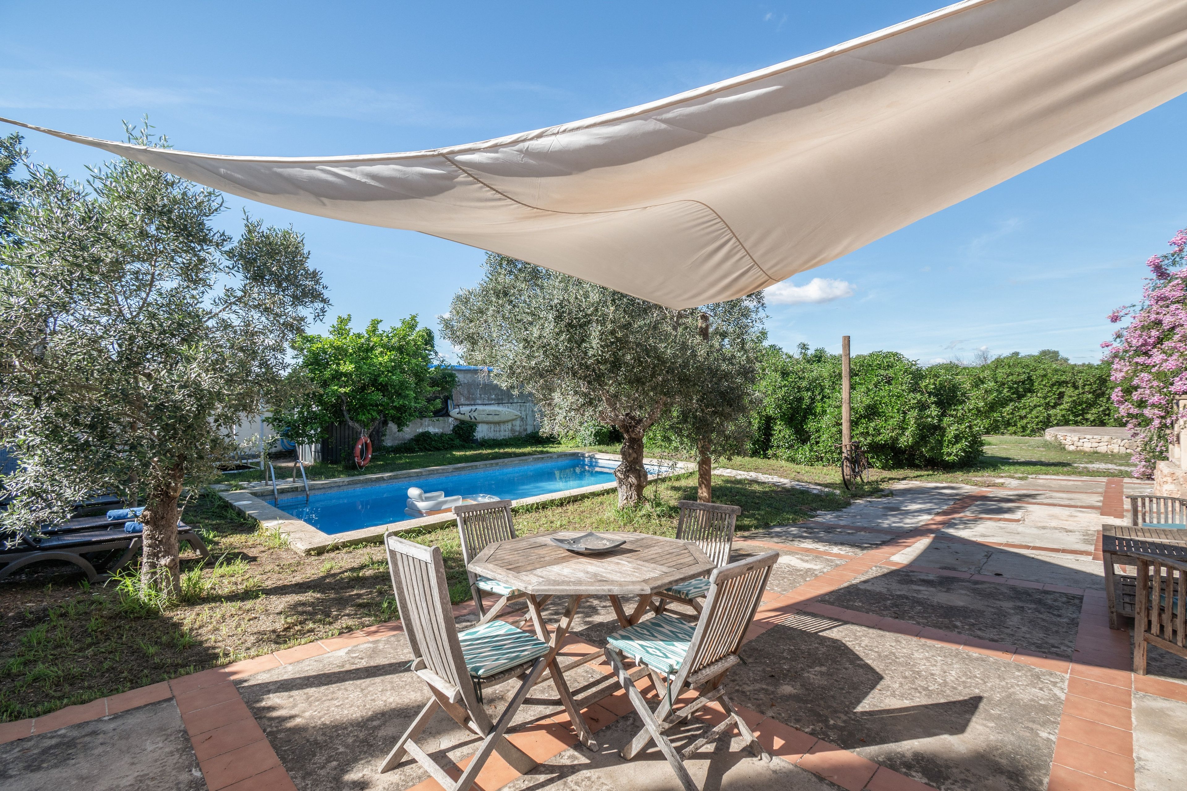 Zona de piscina con mesa y sillas, piscina y abundante vegetación circundante.