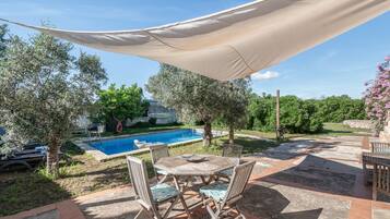 Zona de piscina con mesa y sillas, piscina y abundante vegetación circundante.