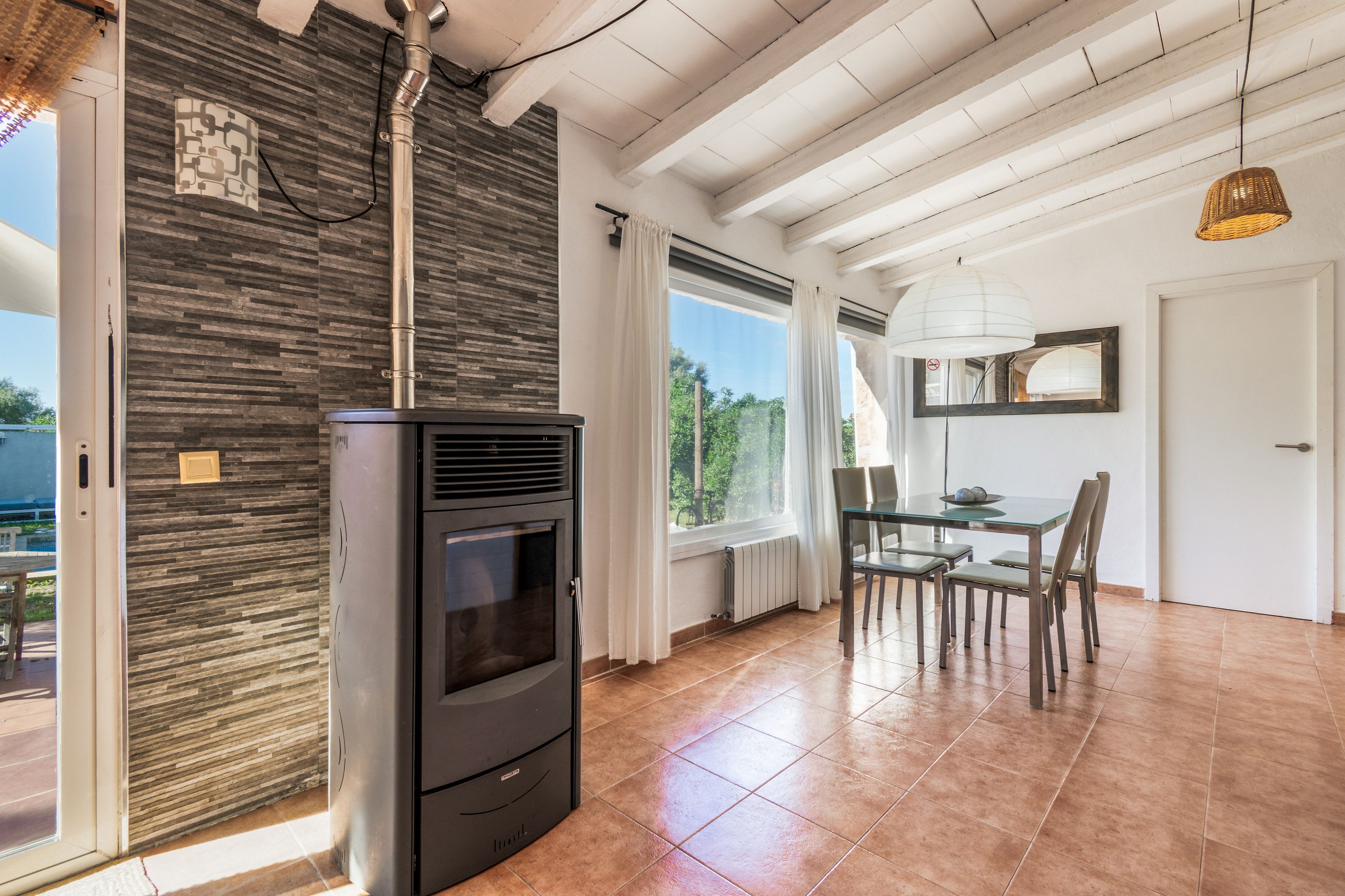 Una cocina moderna con estufa de leña, zona de comedor con mesa y sillas de vidrio, y un amplio ventanal con cortinas.