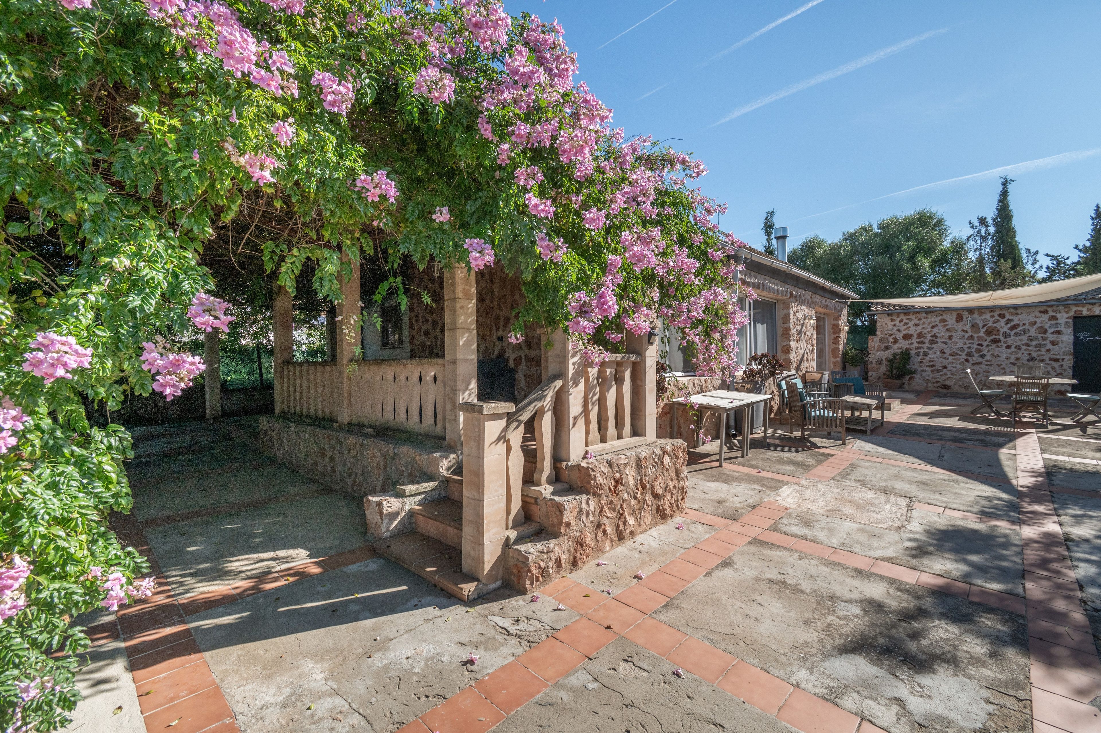 Un edificio de piedra con una pérgola cubierta de flores rosadas, un patio con mobiliario exterior y un cielo azul despejado.