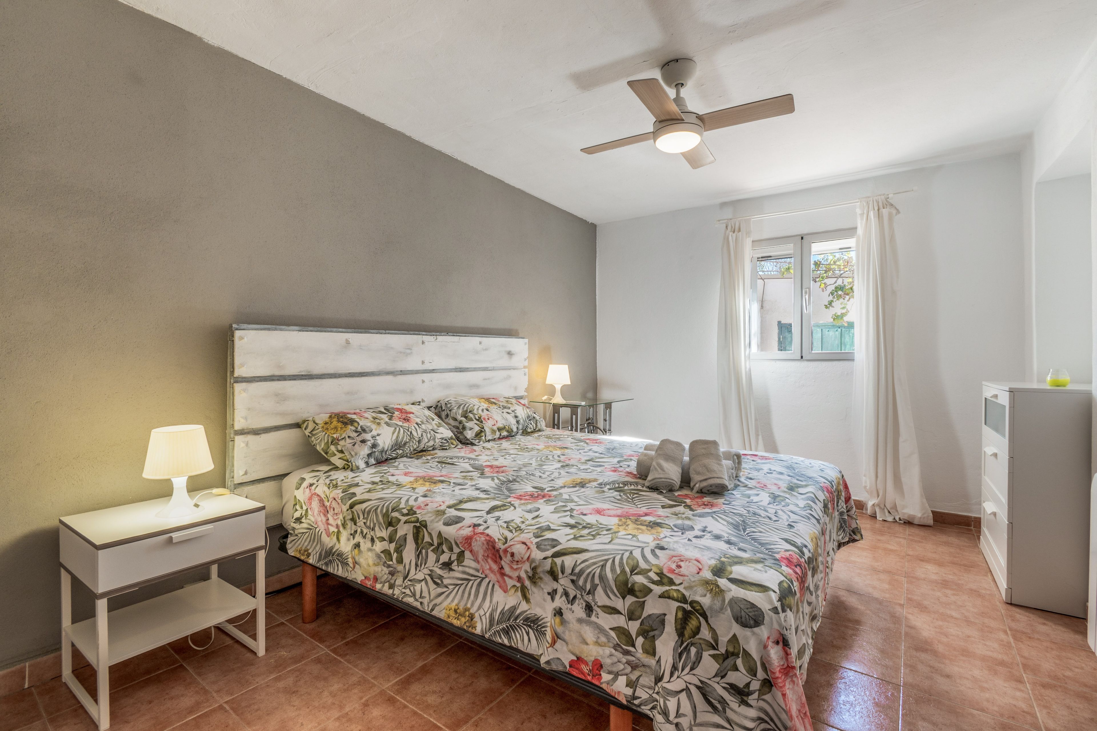 Un dormitorio con cama, mesita de noche, lámpara, ventilador de techo y ventana con cortinas.