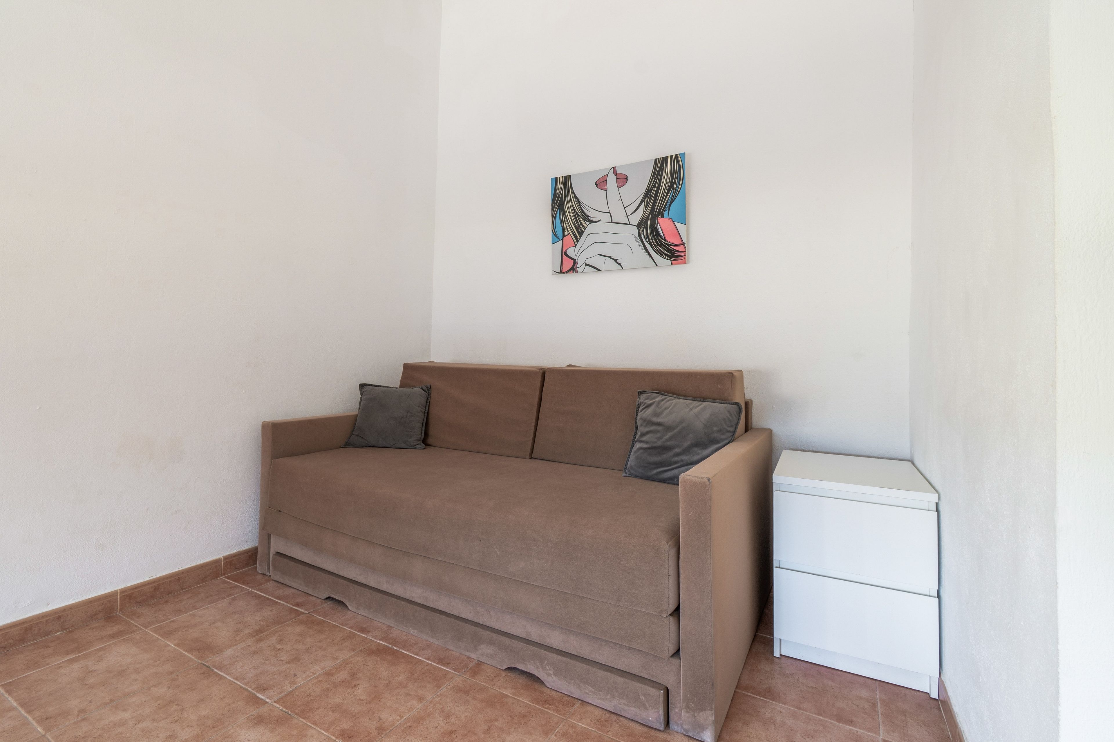 Una stanza con un divano marrone, due cuscini, un piccolo armadietto bianco e un'opera d'arte appesa al muro.