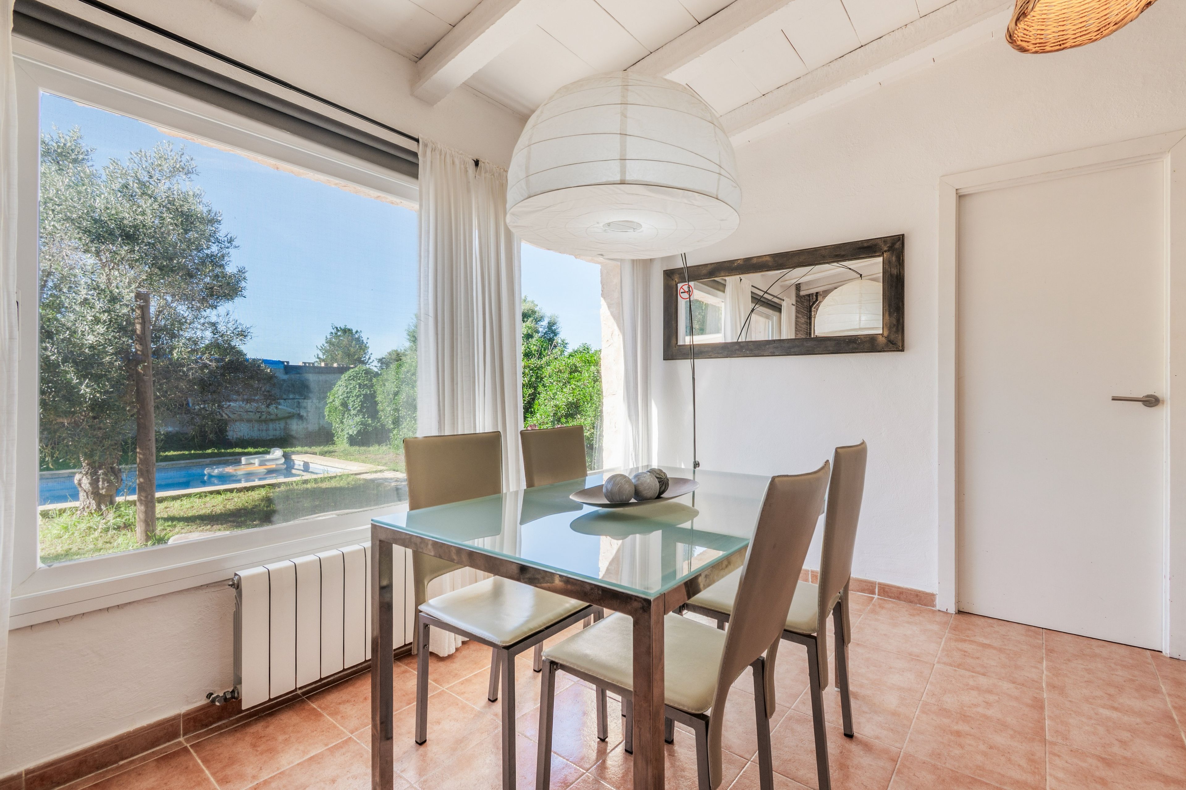 Zona pranzo con tavolo e sedie in vetro, vista su piscina e vegetazione all'esterno, soffitto bianco e una porta.