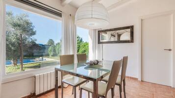 Zona pranzo con tavolo e sedie in vetro, vista su piscina e vegetazione all'esterno, soffitto bianco e una porta.