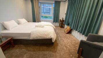 A hotel room with a large bed, a chair, a small table, and a window with curtains.
