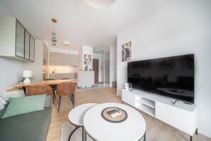 A modern living room with a flat-screen TV, a dining area with a wooden table and chairs, and a kitchen in the background. - Kierbedzia Premium Apartament, Mokotów (Warszawa)