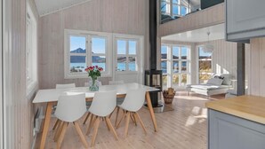 A modern interior with a dining area, white furniture, and large windows offering a view of the outdoors. - Oceanview Cabin Sleeps 6 - Sauna - Private Terrace (Bø)