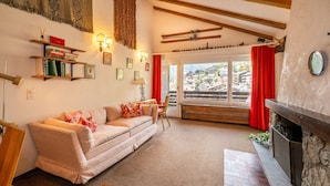 A cozy living room with a sofa, a small table, a fireplace, and a view of a mountain village through a large window.