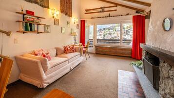 A cozy living room with a sofa, a small table, a fireplace, and a view of a mountain village through a large window.
