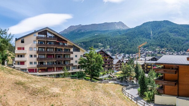 A multi-story hotel with balconies, set against a mountainous backdrop.