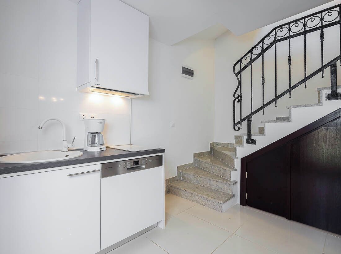 Une cuisine moderne avec des meubles blancs, une machine à café et un lave-vaisselle. Un escalier avec une rampe noire mène à une pièce sombre.