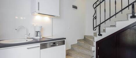 Une cuisine moderne avec des meubles blancs, une machine à café et un lave-vaisselle. Un escalier avec une rampe noire mène à une pièce sombre.
